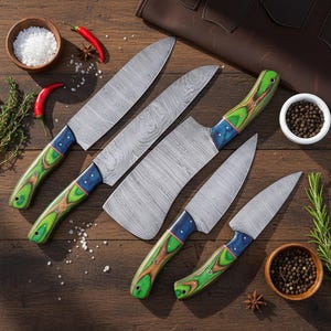 Op de afbeelding: Een set van vijf Damascus stalen keukenmessen met groene, blauwe en bruine houten handvatten. De messen hebben een uniek patroon. De messen liggen op een houten oppervlak met kruiden en specerijen.