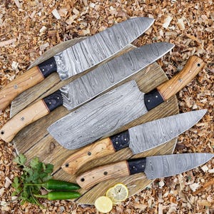 May include: A selection of Damascus steel kitchen knives with wooden handles and black details, arranged on a wooden board. The blades have a distinctive pattern. The image includes green chillies, coriander, and lemon slices, with wood shavings in the background.