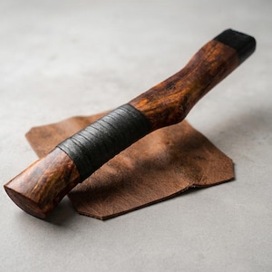 May include: A cylindrical wooden handle with a polished brown grain, featuring a black wrapped grip section in the centre and a solid black cap at the end, resting diagonally across a small, square piece of textured brown leather material.