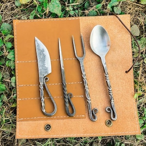 May include: A handcrafted cutlery set with a knife, fork, spoon, and skewer. The utensils have twisted handles and are displayed on a tan leather roll with brass snaps and a leather tie.