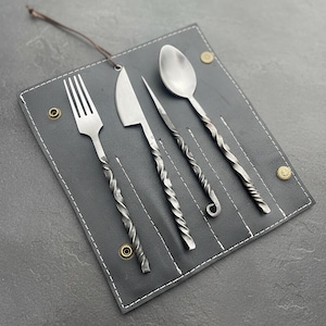May include: A set of four handcrafted metal utensils: a fork, knife, skewer, and spoon, each with a twisted handle design. The utensils are displayed in a dark gray leather roll with white stitching and brass-colored snaps.
