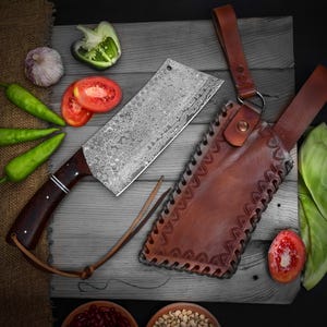 May include: A Damascus steel cleaver with a dark wood handle and leather sheath. The blade has a unique pattern. The sheath is brown with decorative stitching. Fresh vegetables, including tomatoes, peppers, and garlic, are arranged around the knife.