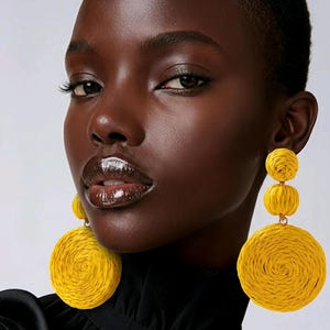 May include: A woman with dark skin wearing large, yellow, woven earrings. The earrings have a circular design with a spiral pattern. The woman is wearing a black top. The background is a light grey.
