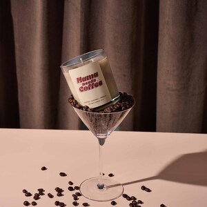 Könnte beinhalten: Eine Kerze in einem Martini-Glas gefüllt mit Kaffeebohnen. Die Kerze hat ein Etikett mit der Aufschrift "Mama needs Coffee". Die Kerze ist beige mit weißem Etikett und rotem Text. Kaffeebohnen sind verstreut.