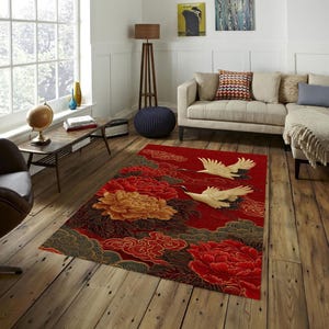 May include: A red area rug with a floral and crane design. The rug features large red and gold peony flowers, green and gold clouds, and two white cranes in flight. The rug is placed on a hardwood floor in a living room setting.