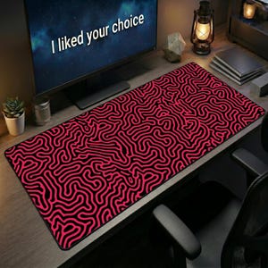 May include: A rectangular desk mat featuring a vibrant pink and black maze-like pattern of interconnected lines. The mat is positioned on a wooden desk surface in front of a computer monitor displaying the text I liked your choice in white.