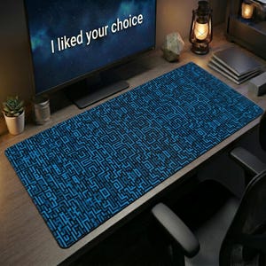 Op de afbeelding: Een rechthoekige bureaumat met een herhalend donkerblauw en zwart doolhofpatroon. De mat ligt op een houten oppervlak voor een computermonitor die de tekst I liked your choice in witte letters weergeeft tegen een donkere achtergrond.