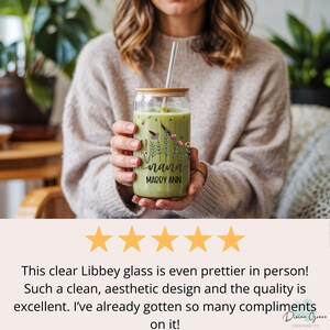 May include: A clear glass tumbler with a bamboo lid and straw, filled with a green beverage. The glass is decorated with floral and insect illustrations, and the name "nana" and "MARRY ANN" are printed on the glass. The tumbler is held by a person.