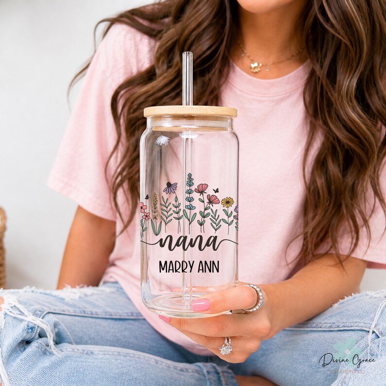 May include: A clear glass can-shaped tumbler with a bamboo lid and a straw. The tumbler features a floral design with the word "nana" and the name "Marry Ann" printed on it. The design includes colorful flowers and greenery.