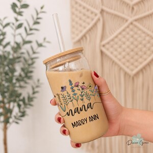 May include: A clear glass tumbler with a wooden lid and straw, filled with a light brown beverage. The tumbler features a floral design with the word "nana" and the name "Marry Ann" in black script. The glass is held by a person.