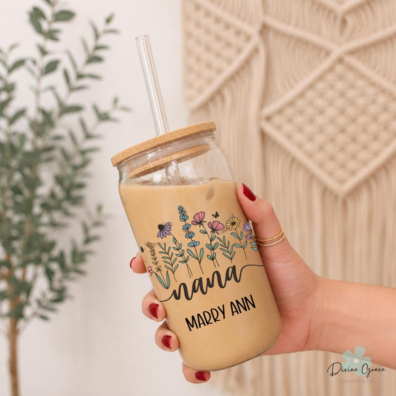 May include: A clear glass tumbler with a wooden lid and straw, filled with a light brown beverage. The tumbler features a floral design with the word "nana" and the name "Marry Ann" in black script. The glass is held by a person.
