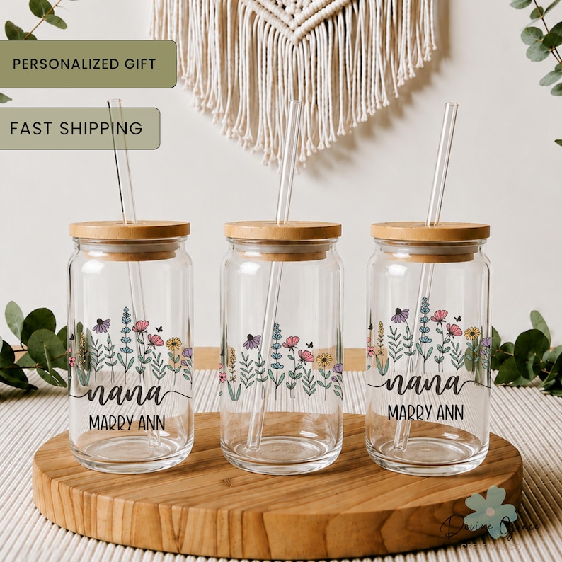 May include: Three clear glass drinking jars with bamboo lids and glass straws. Each jar features a floral design and the name "nana" with "Marry Ann" underneath. The jars are on a wooden tray.