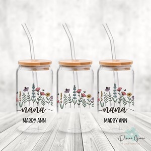 May include: Three clear glass drinking jars with wooden lids and glass straws. Each jar features a floral design with the word "nana" and the name "Marry Ann" in black script. The jars are decorated with colorful flowers and butterflies.