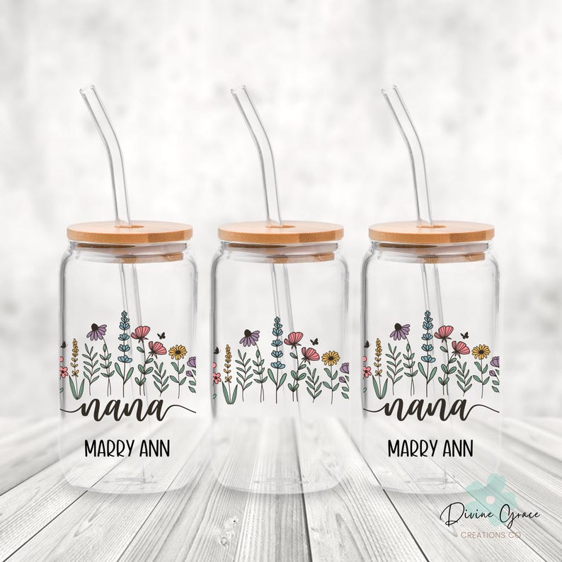 May include: Three clear glass drinking jars with wooden lids and glass straws. Each jar features a floral design with the word "nana" and the name "Marry Ann" in black script. The jars are decorated with colorful flowers and butterflies.