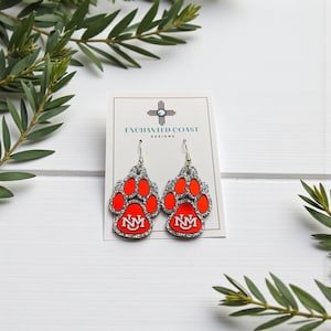 May include: A pair of paw-shaped earrings with a silver glitter outline and bright orange pads. Each earring features the letters "NM" in white on a red background. The earrings hang from silver hooks, and are displayed on a white card.
