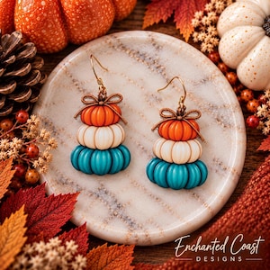 May include: A pair of dangle earrings featuring a stacked design of three pumpkins in orange, white, and teal. Each earring is topped with a small brown bow and attached to a gold-toned hook. The earrings are displayed on a round marble surface.
