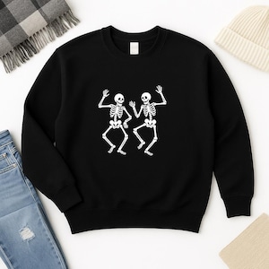 Puede incluir: Sudadera negra con un gráfico blanco de dos esqueletos bailando. Los esqueletos se representan con estructuras óseas detalladas y brazos levantados, creando un diseño juguetón. La sudadera es de estilo cuello redondo clásico.