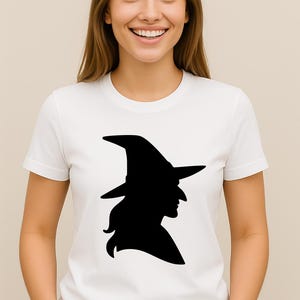 May include: White t-shirt featuring a black silhouette of a witch wearing a pointed hat. The graphic is centered on the chest. The model is wearing a white t-shirt.