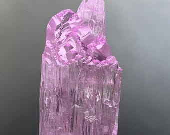 May include: A translucent, light purple crystal formation. The crystal has a columnar structure with vertical striations and a jagged, crystalline top. The base is mounted on a small, rectangular stand. The crystal's color is a soft, pastel purple.