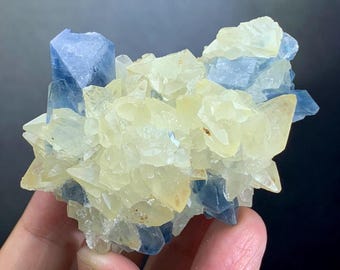 May include: A cluster of crystals featuring a mix of pale yellow and blue hues. The crystals have a rough, textured appearance, with sharp edges and varying sizes. The overall shape is irregular and organic, with a natural, earthy aesthetic.