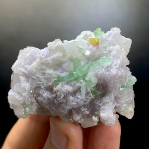 May include: A close-up of a mineral specimen featuring a cluster of crystals. The specimen has a light purple base with clear and green crystal formations. A small, yellow crystal is visible on top.