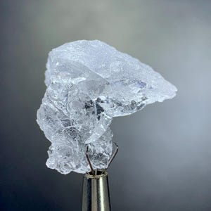 Può includere: Un campione di minerale cristallino trasparente, tenuto da uno strumento di metallo. Il minerale ha una superficie ruvida e strutturata e appare traslucido. Lo sfondo è un gradiente di grigio e bianco.