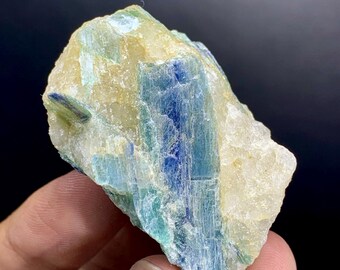 May include: A raw, natural kyanite crystal with a mix of blue, teal, and cream colors. The crystal has a layered, striated appearance, with the blue hues concentrated in the center. The stone is held in a hand against a dark background.