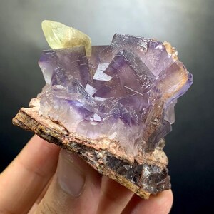 Puede incluir: Un grupo de cristales de fluorita c&uacute;bicos y transl&uacute;cidos de color p&uacute;rpura unidos a una matriz mineral rugosa de color gris parduzco. Un cristal &uacute;nico, alargado y de color amarillo p&aacute;lido, sobresale del lado superior izquierdo.