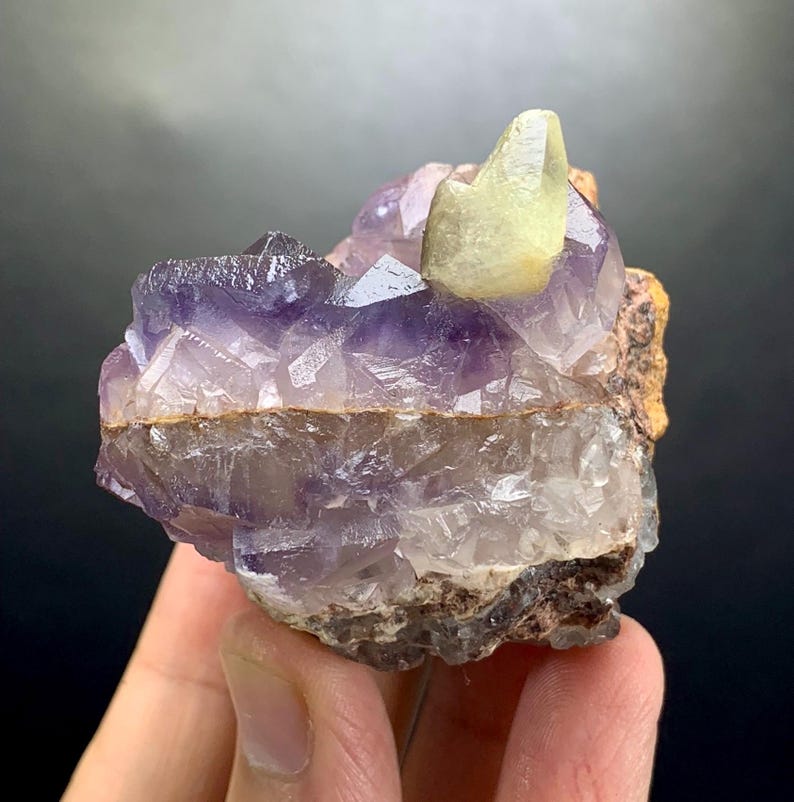 Puede incluir: Un esp&eacute;cimen mineral en bruto con cristales de amatista de color p&uacute;rpura intenso agrupados. Un cristal de calcita de color amarillo p&aacute;lido y liso se asienta de forma prominente sobre la formaci&oacute;n p&uacute;rpura, unida a una base de roca marr&oacute;n, rugosa y terrosa.