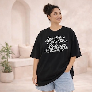 May include: Black oversized t-shirt with the text "Gotta Keep An Eye Out For Selener" in white script. The shirt has short sleeves and a crew neck. The model is wearing blue denim shorts.