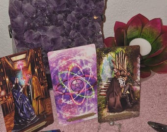 May include: A collection of tarot cards, a chakra pendant, and an amethyst crystal cluster are displayed. The tarot cards depict illustrations of figures and symbols. A red and green lotus flower candle holder is also visible.