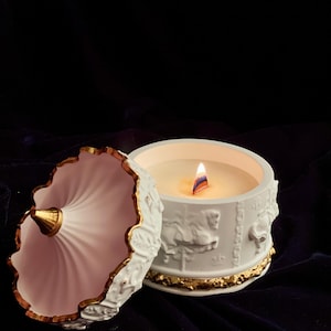Op de afbeelding: Een brandende witte kaars in een decoratieve container met een deksel. De container is wit met gouden randen en reliëfdetails van carrouselpaarden. Het deksel is roze met gouden randen.