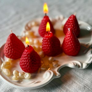 May include: Six red strawberry-shaped candles, some lit with yellow flames, are displayed on a white, ornate plate. The candles have a textured surface, resembling a strawberry. Decorative pearls are scattered around the candles.