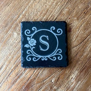 Può includere: Sottobicchiere quadrato in ardesia nera con un motivo floreale e a volute decorative. Il design presenta la lettera "S" in una cornice circolare. Il sottobicchiere è posto su una superficie di legno.