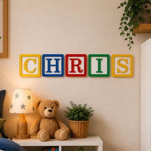 May include: Five square wall-mounted letter boards arranged horizontally to spell the name CHRIS. Each board features a white grooved background with a coloured frame and a large, bold, contrasting letter in the centre: yellow C, blue H, red R, green I, and yellow S.