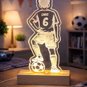 May include: A glowing LED night light shaped like a soccer player, standing on a soccer ball. The player's jersey has the name "CHRIS" and the number "6". The light sits on a rectangular wooden base.