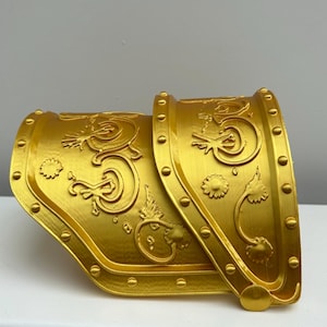 May include: A gold-coloured arm guard with ornate, raised floral and scrollwork designs. The metal armour features a curved shape with rivets along the edges, likely part of a costume or historical reenactment ensemble.