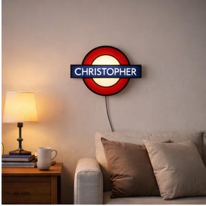 May include: A wall-mounted light fixture in the style of a London Underground sign, with a red circle and a blue rectangular bar displaying the name "CHRISTOPHER" in white. The fixture is illuminated, casting a warm glow.