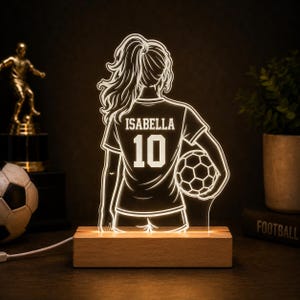 May include: An illuminated acrylic lamp featuring a line-art silhouette of a football player from behind, wearing a jersey with the name ISABELLA and the number 10, holding a football, mounted on a rectangular light-coloured wooden base.