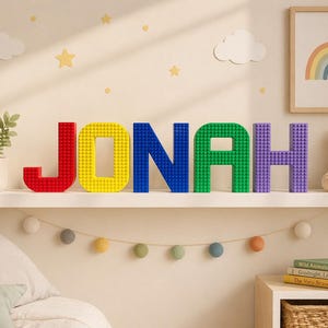 Op de afbeelding: Op een plank staat de naam "JONAH" in kleurrijke letters in blokstijl. De letters zijn rood, geel, blauw, groen en paars. Een teddybeer en een ingelijste regenboogafbeelding staan ook op de plank. Een kleine plant en decoratieve lampjes zijn ook zichtbaar.