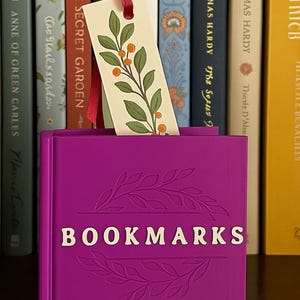 Può includere: Un porta segnalibro viola con la scritta "BOOKMARKS" in lettere bianche. Un segnalibro decorativo con un nastro rosso e un design botanico è inserito. Libri sullo sfondo.