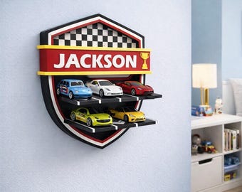 Puede incluir: Estantería de pared para coches con forma de escudo, con el nombre "JACKSON" en letras blancas. La estantería tiene tres niveles para guardar coches de juguete de varios colores. La sección superior tiene un diseño de bandera a cuadros.