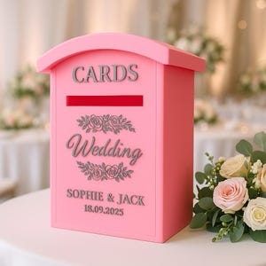 May include: A pink wedding card box with the word "CARDS" at the top. The box has a slot for cards and the word "Wedding" with floral accents. The names "Sophie & Jack" and the date "18.09.2025" are also displayed.