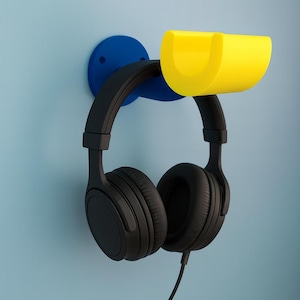 May include: A wall-mounted headphone holder in blue and yellow, holding a pair of black headphones. The holder has a circular blue base and a curved yellow cradle. The headphones are black with a black cable.