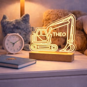 May include: A neon excavator-shaped night light with the name "THEO" in white. The light is on a wooden base, placed on a white bedside table. A blue book and a white alarm clock are also on the table.