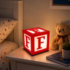 May include: A square, illuminated lamp shaped like a child's alphabet block. The lamp features a bright red frame with a white, glowing centre panel on each visible side. A large, bold, red letter F is displayed on the front and top faces.