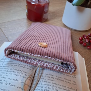 Può includere: Una custodia per libri in velluto a coste color rosa polvere con chiusura a bottone in legno chiaro e un laccetto. La fodera interna presenta un motivo ripetuto a ventaglio in tonalità bianco e rosso spento, appoggiata su un libro aperto.