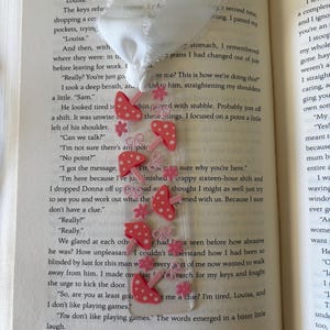 May include: A clear rectangular bookmark featuring a repeating pattern of pink mushrooms with white spots, small pink flowers, and pink bows. The bookmark is topped with a white fabric ribbon tied in a knot at the top edge of the clear plastic.