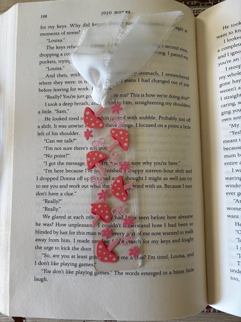 May include: A clear rectangular bookmark featuring a repeating pattern of pink mushrooms with white spots, small pink flowers, and pink bows. The bookmark is topped with a white fabric ribbon tied in a knot at the top edge of the clear plastic.