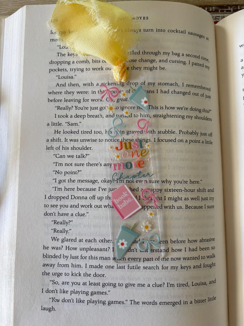 May include: A clear rectangular bookmark with a yellow fabric tassel, featuring colorful graphics of iced coffee cups, pink bows, small white daisies, and a pink book icon. The text "Just one more Chapter" and "book babe" is printed in the center.